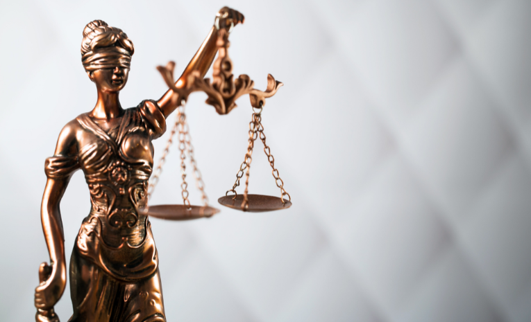 Defensa Legal: Imagen con fondo blanco, rombos sobresaliendo y una estatua dorada de una mujer con los ojos vendados, una espada en la mano izquierda y una balanza en la mano derecha.
