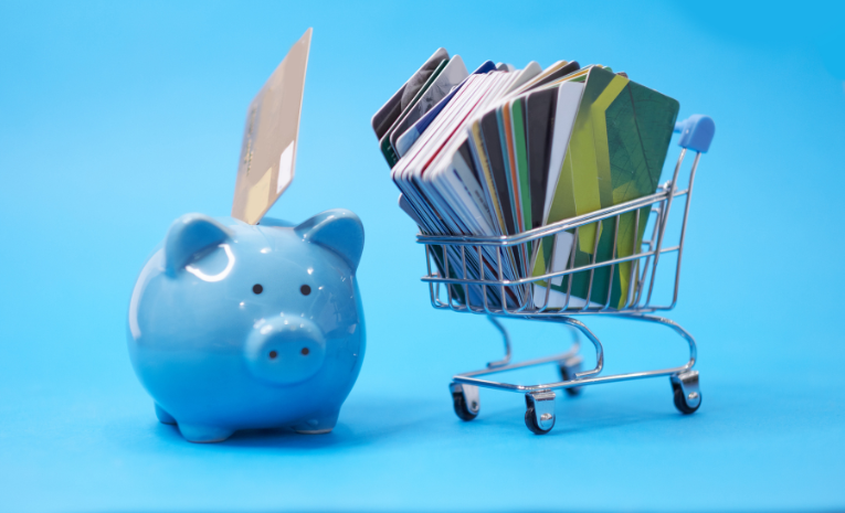 Reestructuración Financiera: Imagen con fondo azul cielo, un carrito de la compra lleno de tarjetas de crédito a la izquierda, y una hucha de cerdito azul con una tarjeta colocada en el hueco de arriba a la derecha.