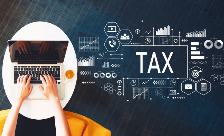 Tax Offices: Imagen de una mesa redonda con un portátil y brazos sobre el teclado, una taza de café junto al portátil, y gráficos y formas con la palabra 'TAX' a la derecha.