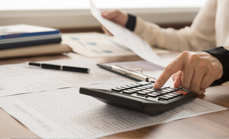 Persona revisando documentos financieros y utilizando una calculadora sobre un escritorio lleno de gráficos y papeles de contabilidad