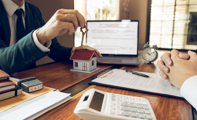 Un agente inmobiliario entregando las llaves de una casa a un cliente en una oficina con documentos y una maqueta de casa sobre la mesa
